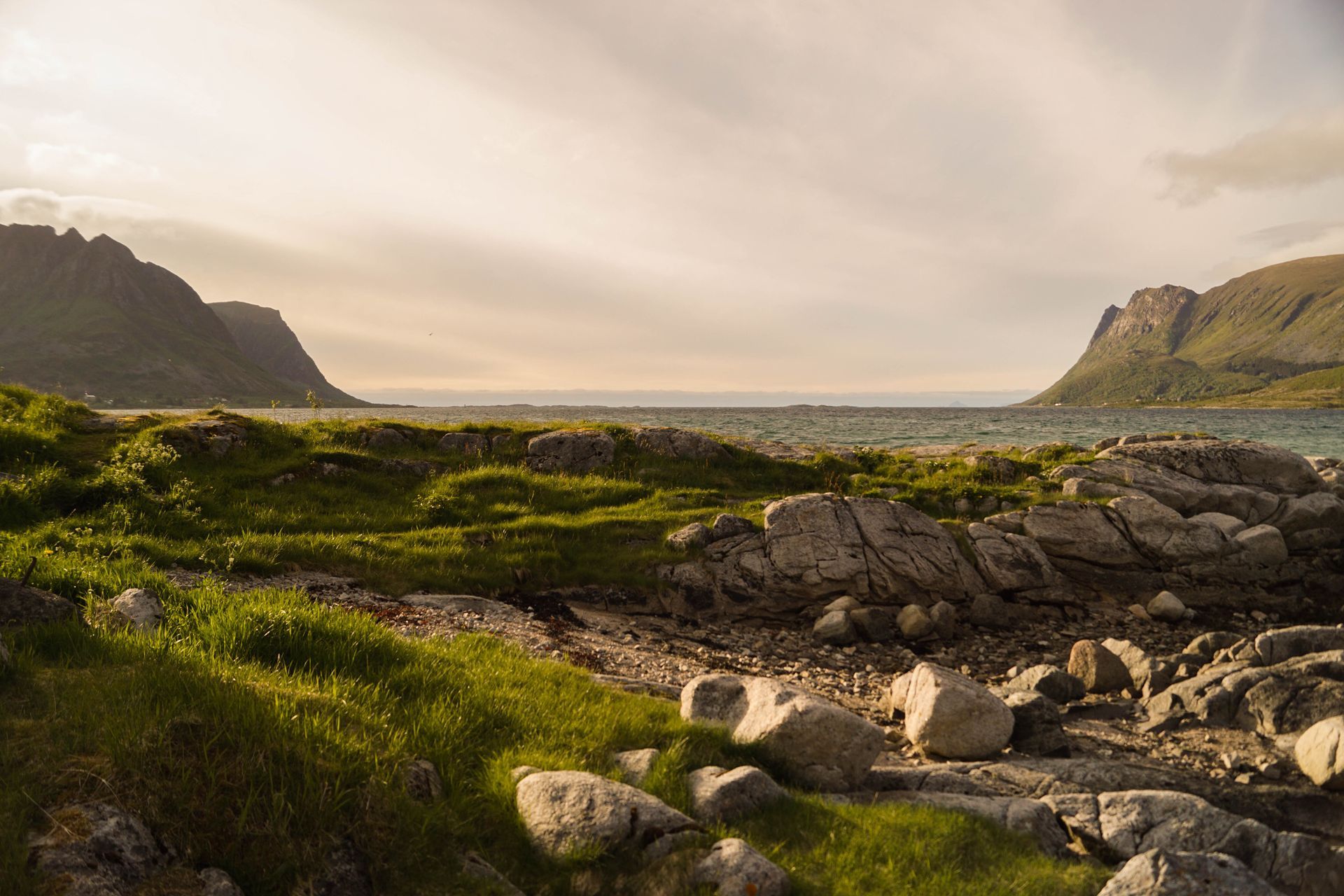 Rocky coast with green grass, mountains, and a body of water under a cloudy sky.