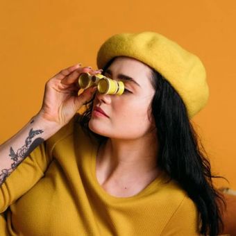 Woman in yellow beret and sweater, looking through yellow binoculars.