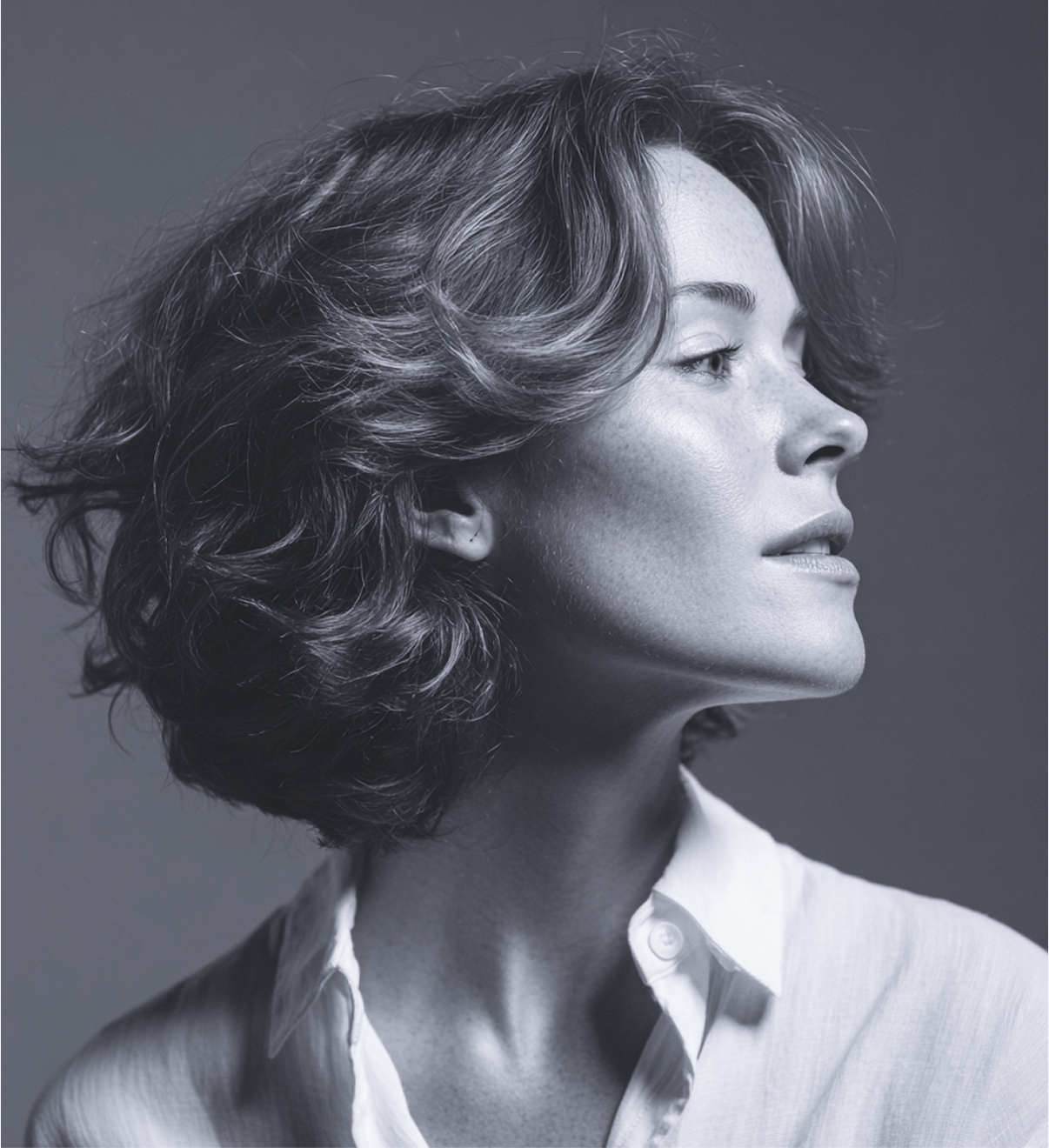 A profile portrait of a person with wavy, shoulder-length hair, wearing a white shirt against a neutral gray background.