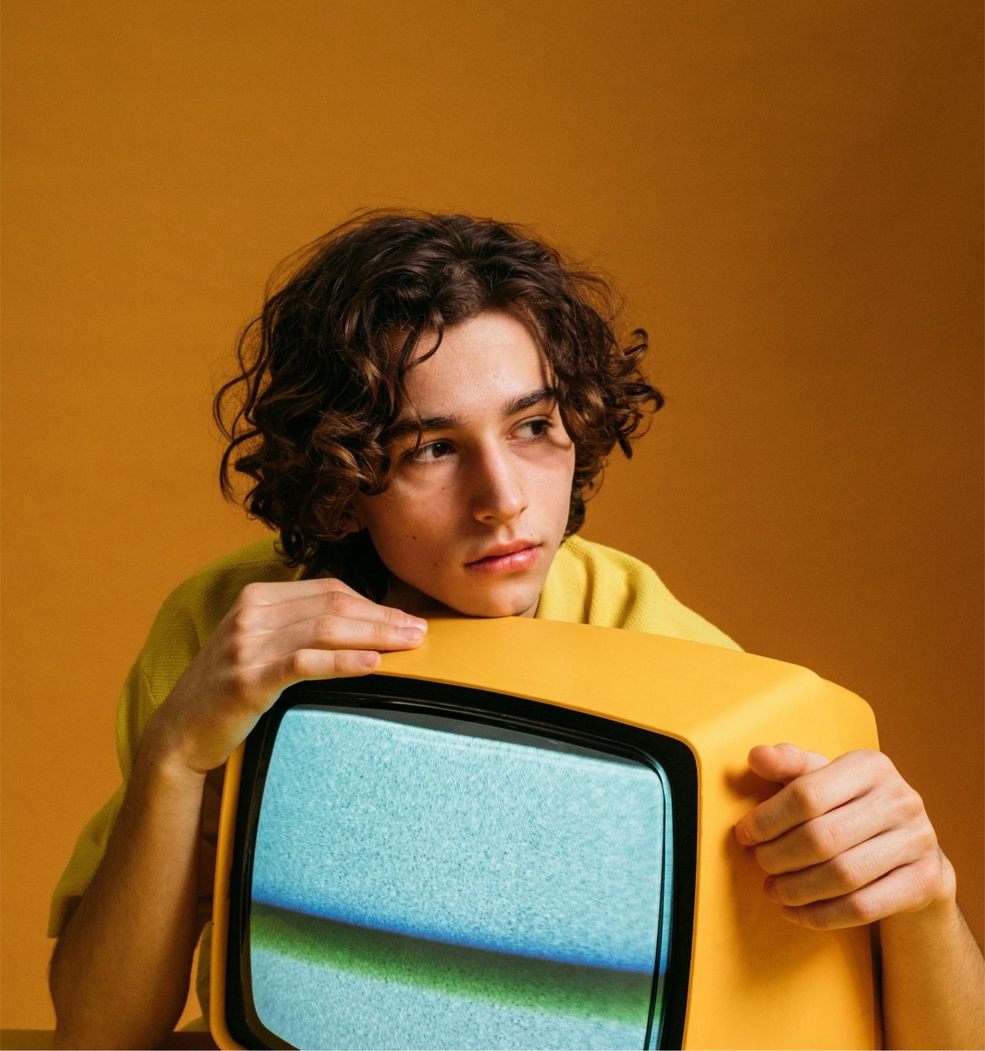 Young person with curly hair rests chin on a yellow, vintage television with static screen. Orange background.