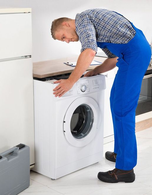 Person in blue overalls fixing a washing machine in a kitchen.