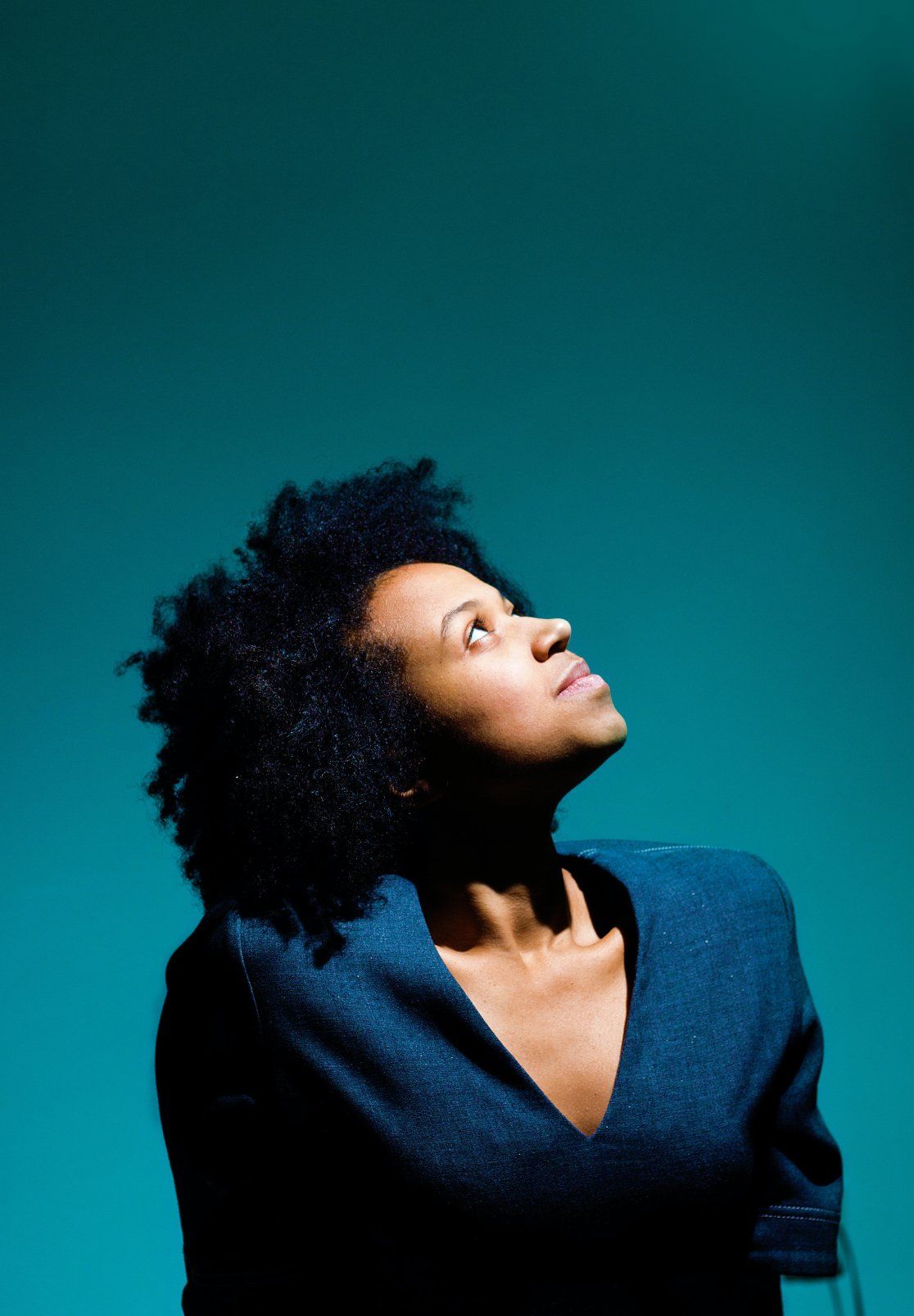 Person with curly hair looking upward against a teal background, wearing a dark blue top.