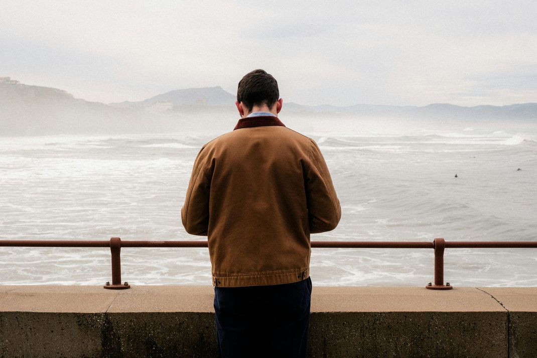 A man in a brown jacket is standing on a wall overlooking the ocean.