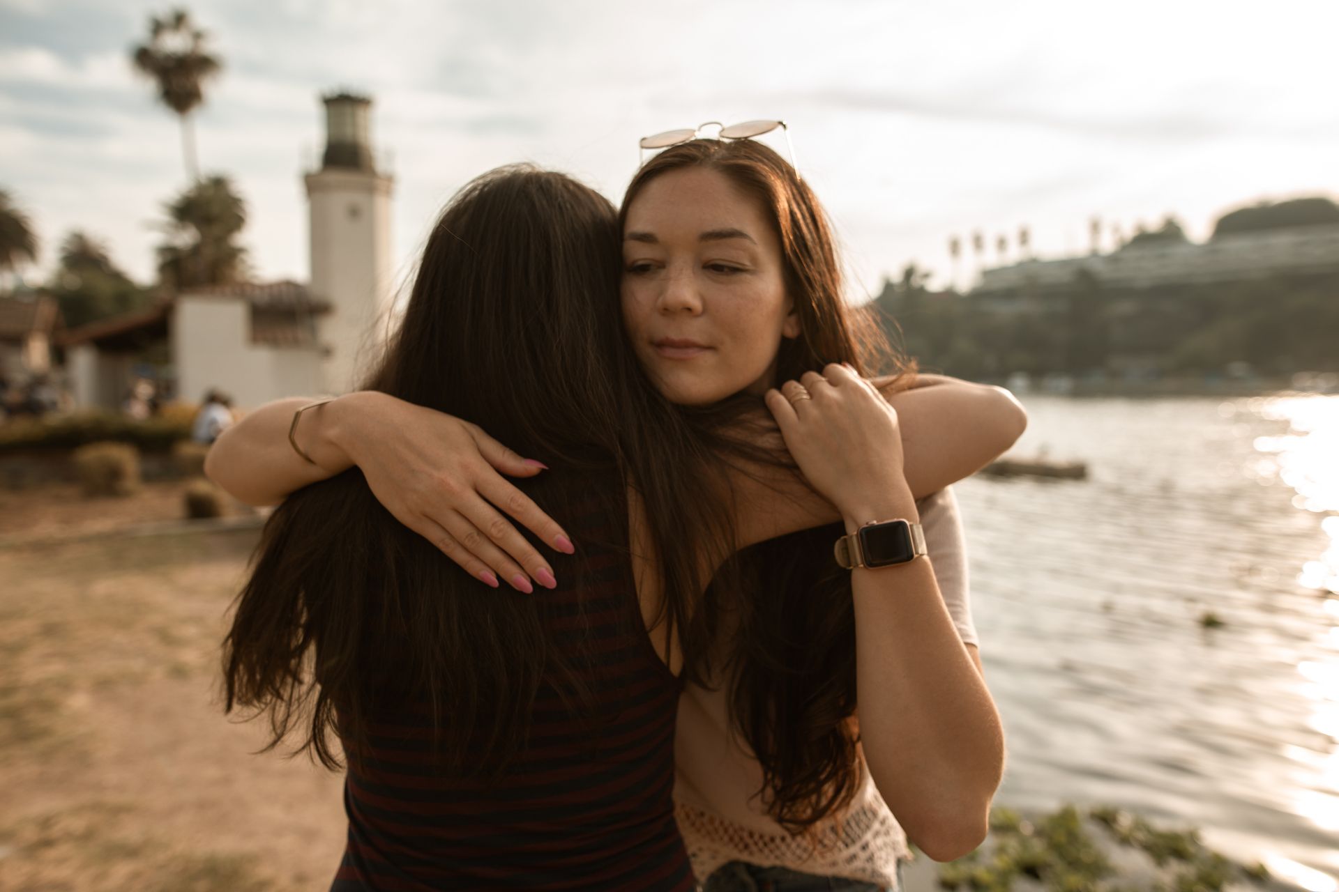 Two women embracing warmly outdoors by a waterfront. One has arms around the other's back, expressing affection and support.