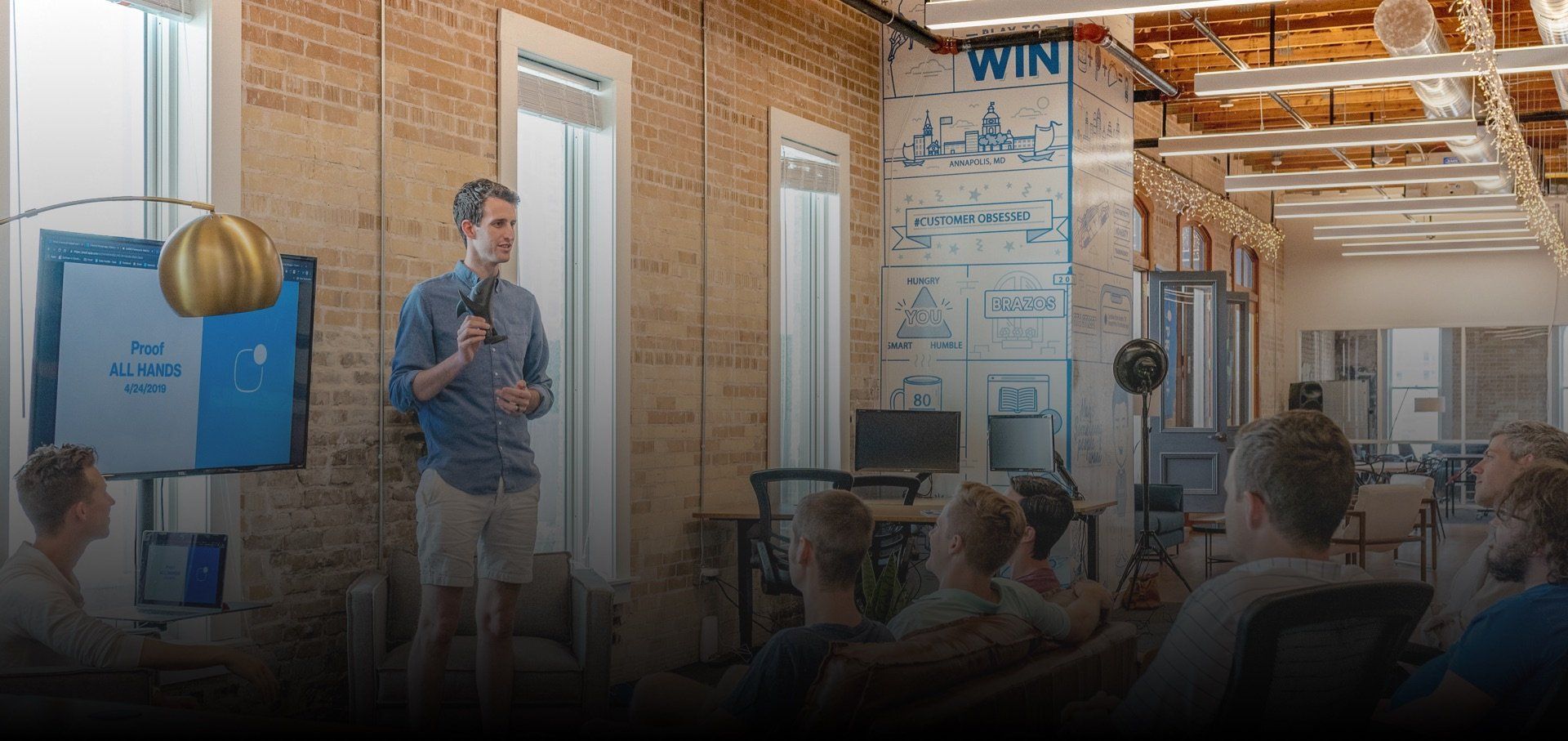 A man gives a presentation to a group of people in a room with brick walls.