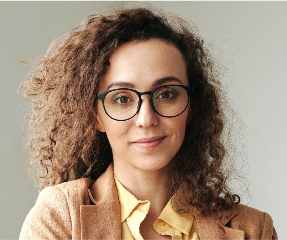A woman with curly hair is wearing glasses and a tan jacket.