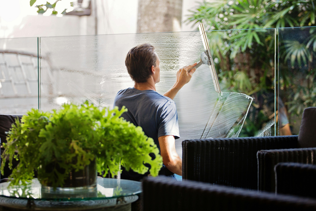 Man squeegeeing a glass window in outdoor setting, with green plants and black wicker furniture.