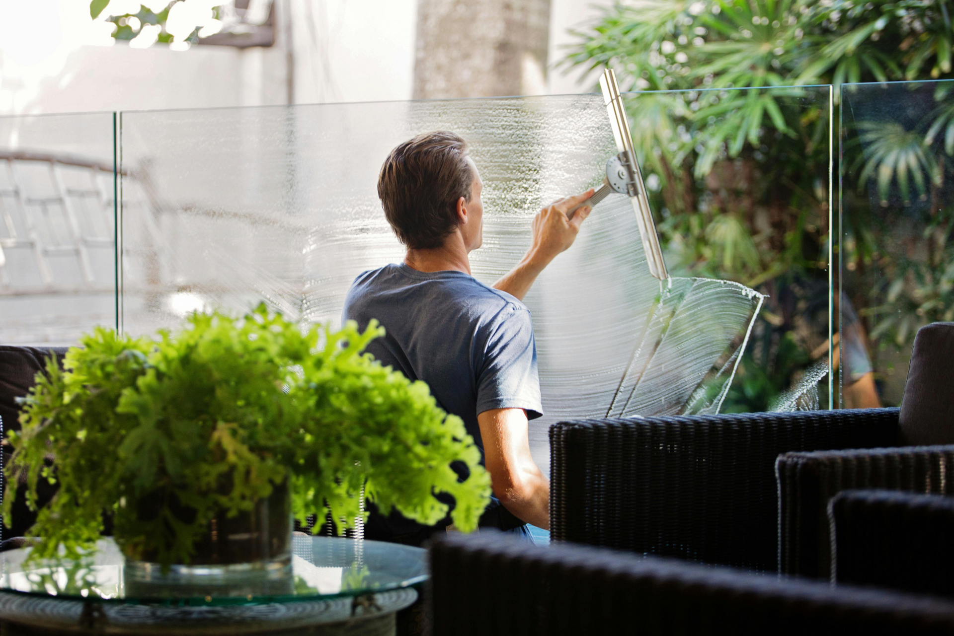 Man squeegeeing a glass window in outdoor setting, with green plants and black wicker furniture.