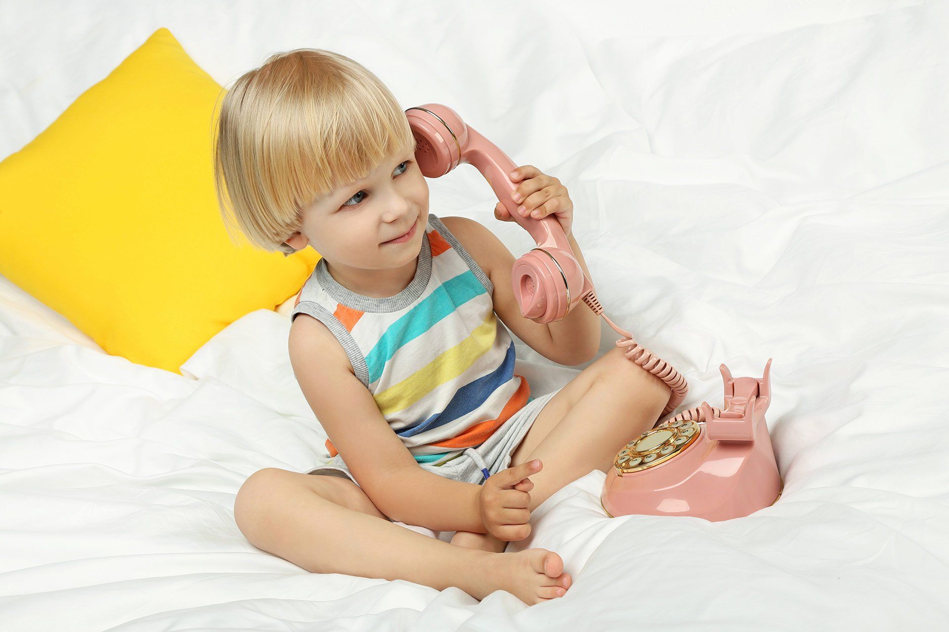 A little girl is sitting on a bed talking on a pink telephone.