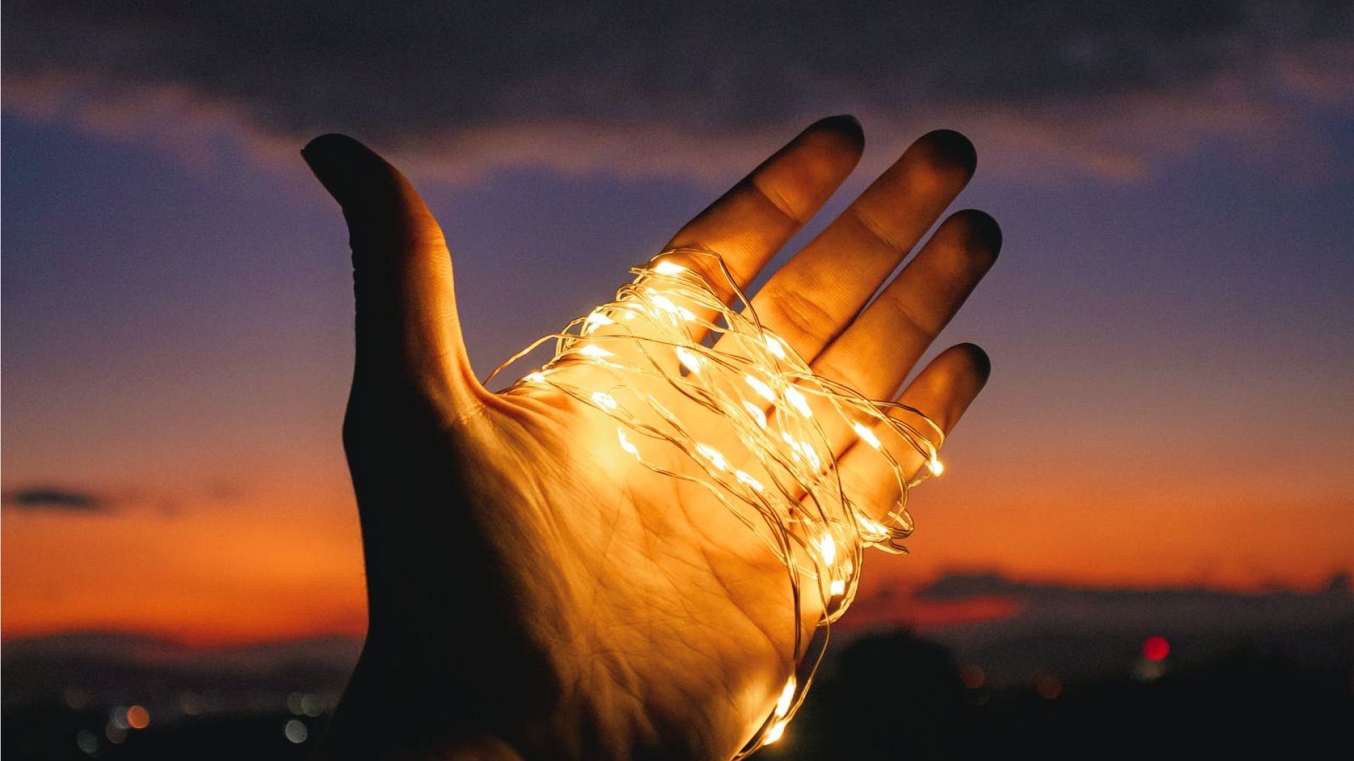 Hand holding string lights against a sunset sky.