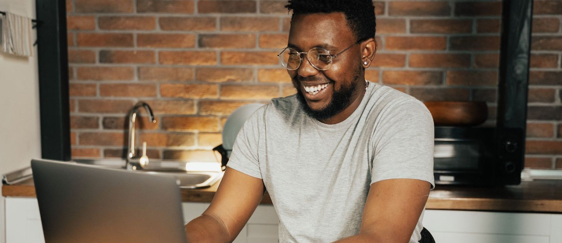 A smiling person with glasses working on a laptop in a kitchen with a brick wall.