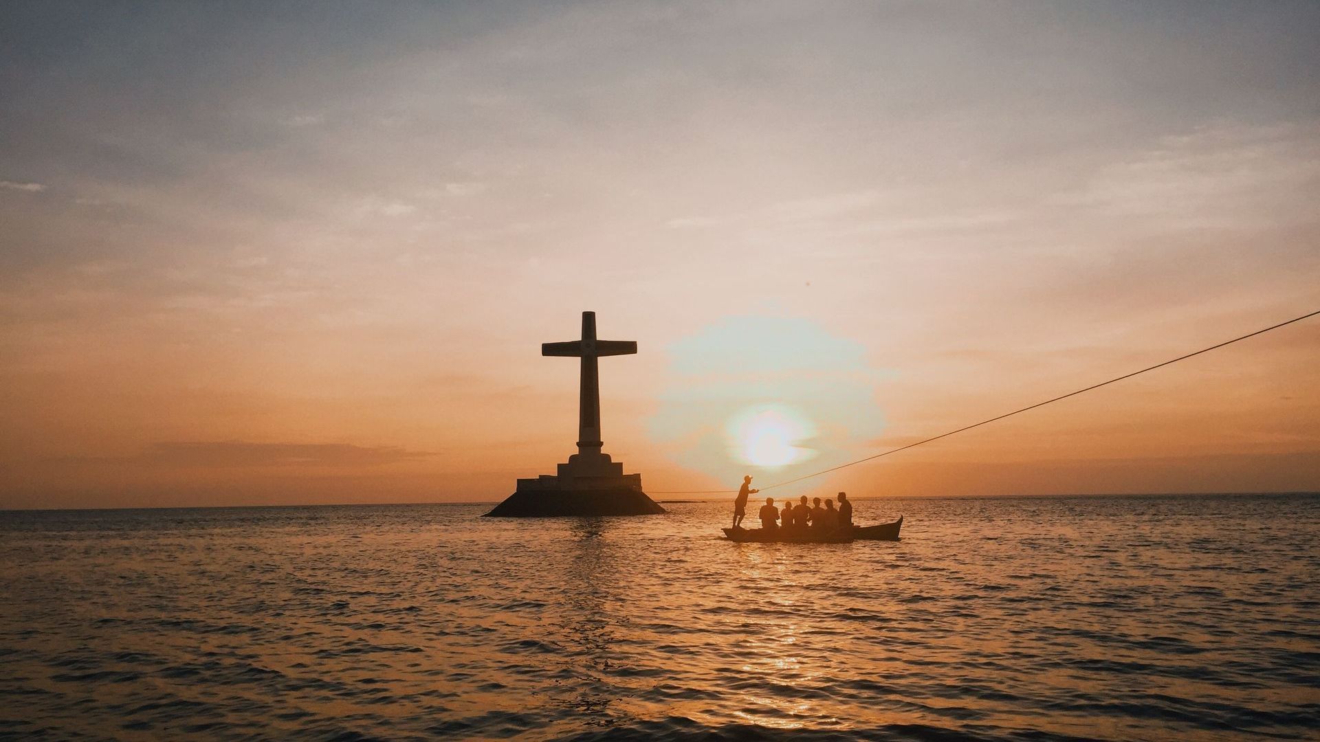 Sunset over ocean with large cross on platform and boat silhouetted against sun.