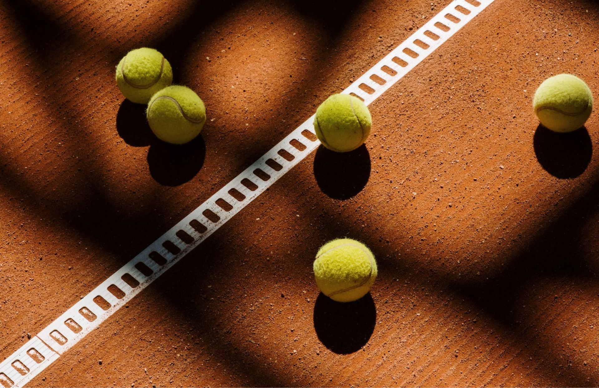Four tennis balls on a clay court beside a white measuring tape in sunlight