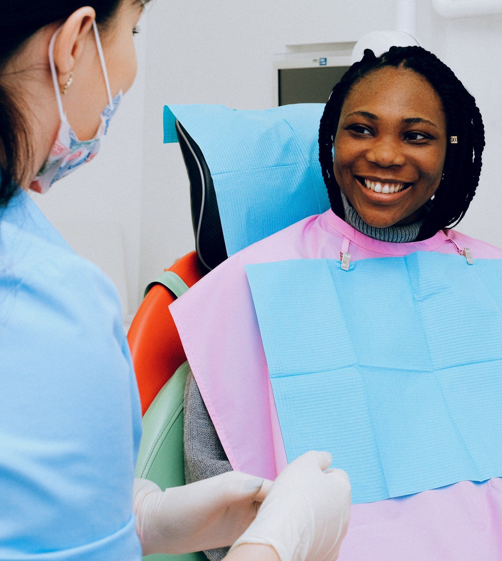 Dentist in blue scrubs examining a smiling Black woman patient in a dental chair; both are wearing masks.
