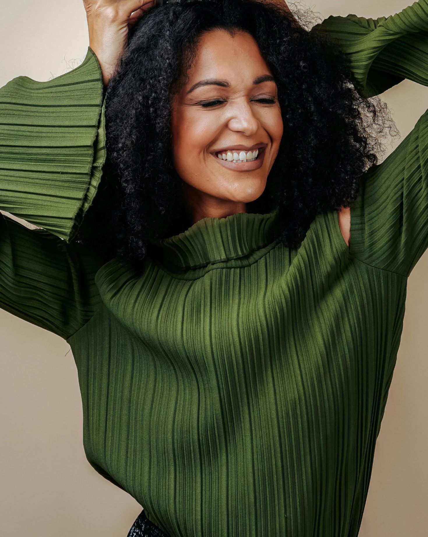 Kvinne med krøllete svart hår smiler, iført en grønn teksturert topp, armene hevet og hendene i håret.