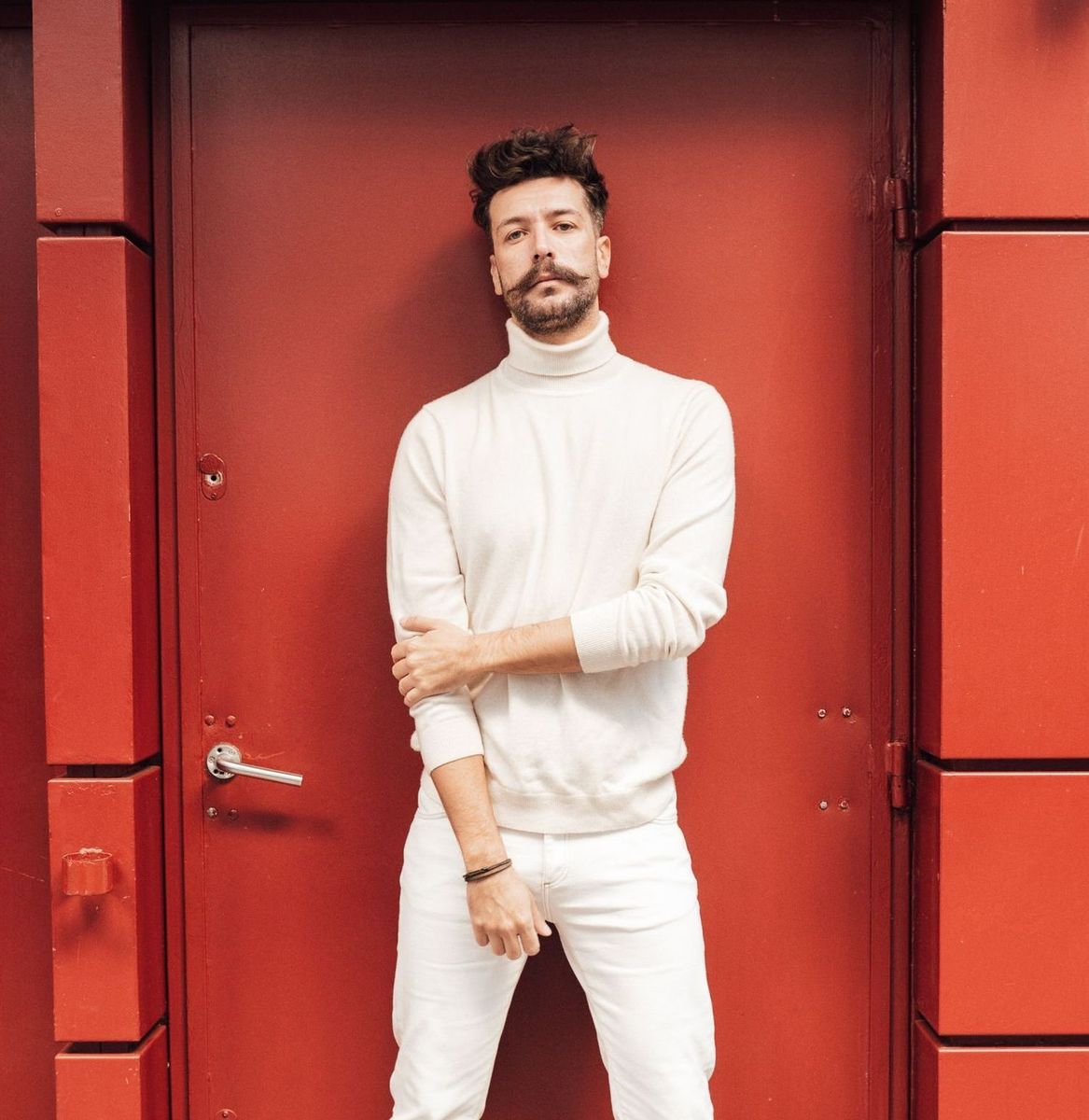 A man in a white sweater and white pants is standing in front of a red door.
