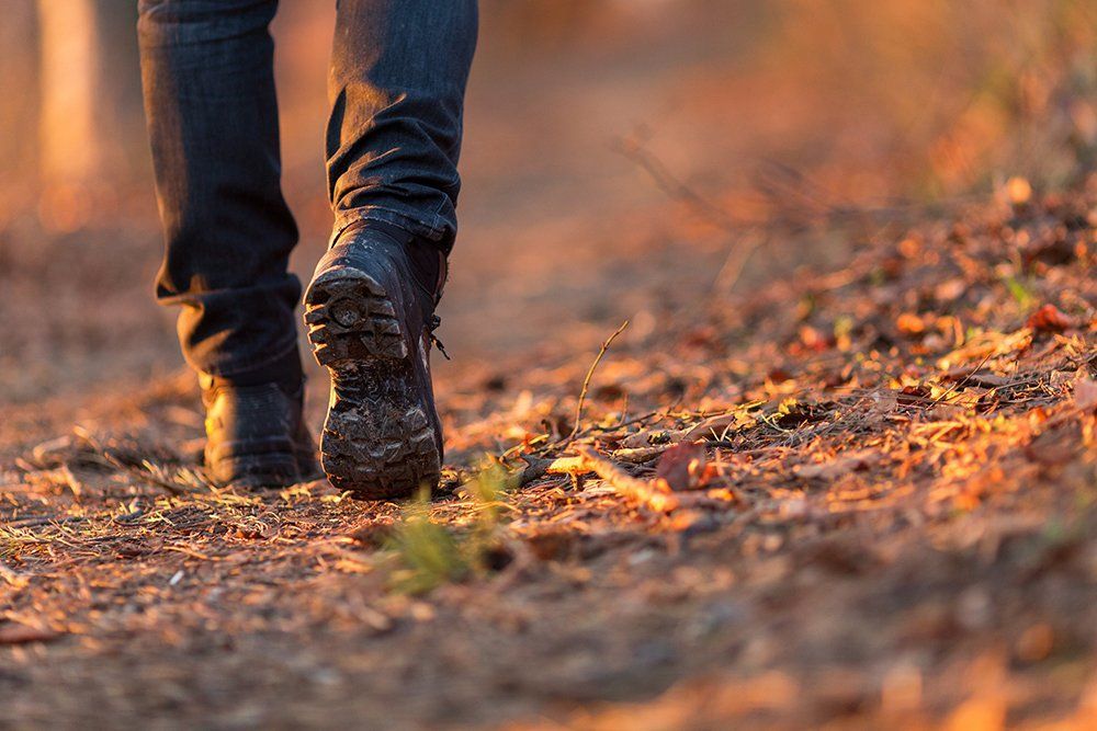 De benen van een persoon in een donkere spijkerbroek en wandelschoenen lopen over een zandpad, in het zonlicht.