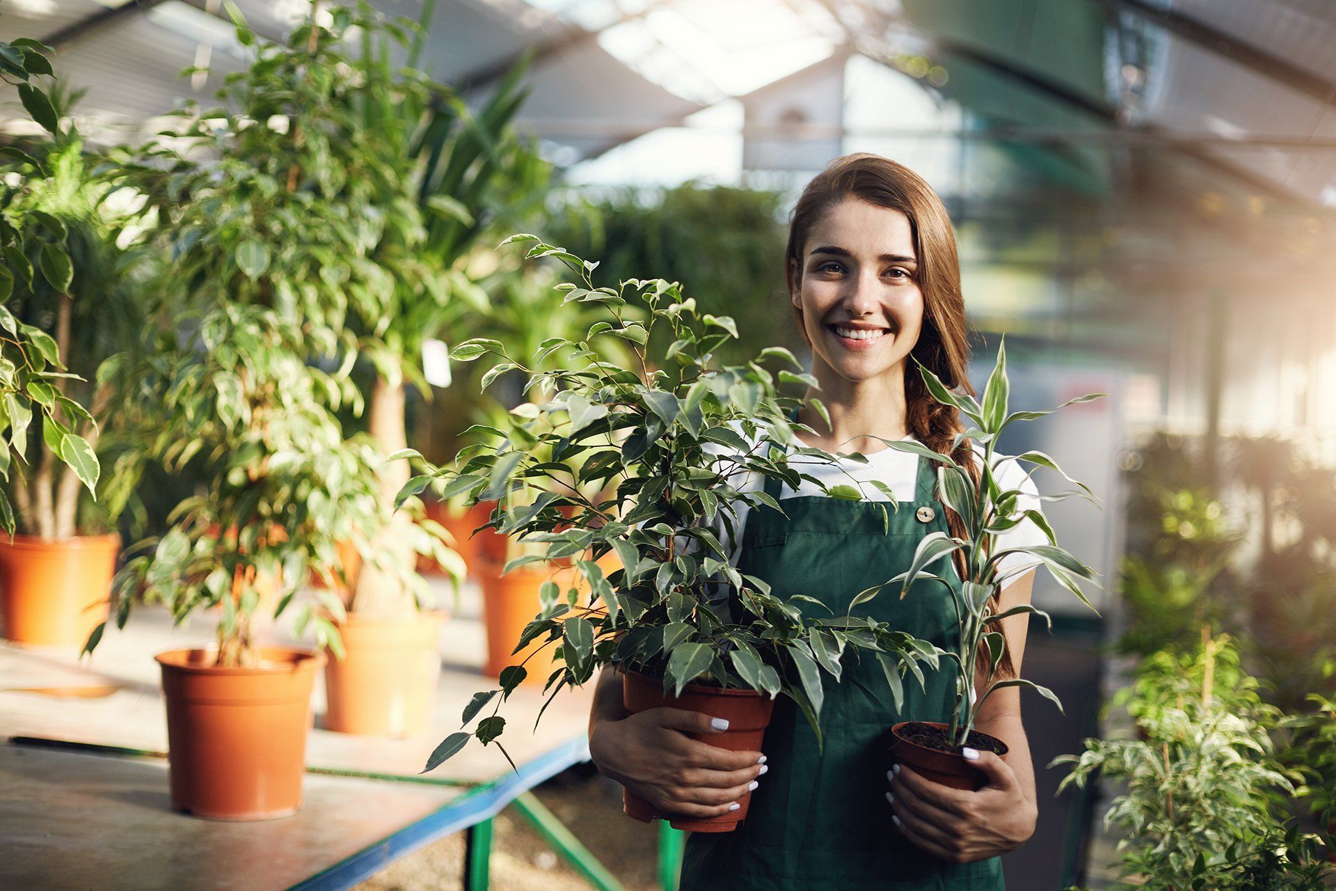 Woman in green apron smiles, holding potted plants in a bright greenhouse.