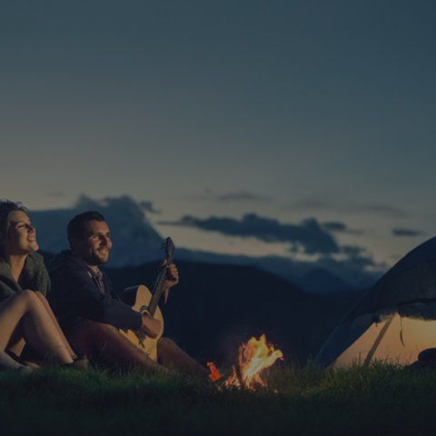Casal acampando; homem tocando violão perto de uma fogueira e barraca sob o céu estrelado.
