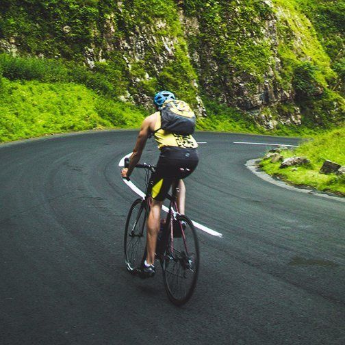 Cyclist in yellow and black riding on a curving road in a green, mountainous landscape.