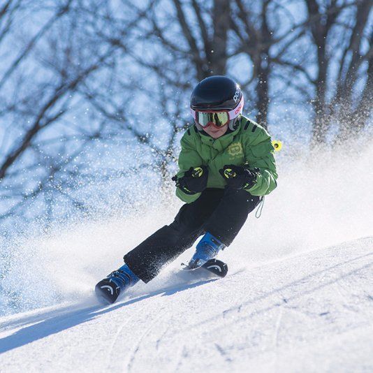 Child skiing down a snow-covered slope wearing a green jacket, helmet, and goggles, kicking up snow.