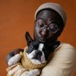 A person wearing a beanie and glasses holding and gently kissing a small, black-and-white dog in a tan sweater.