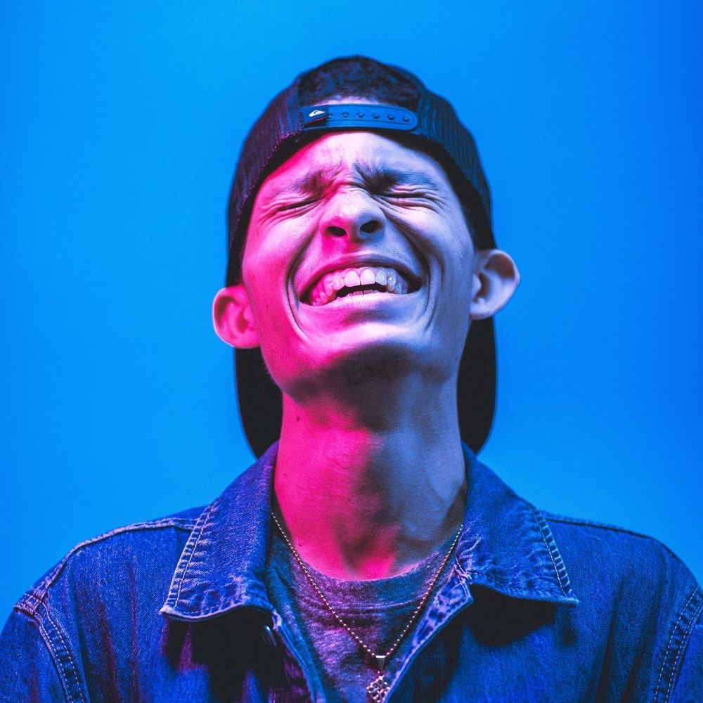 A man wearing a hat and a denim jacket is laughing.