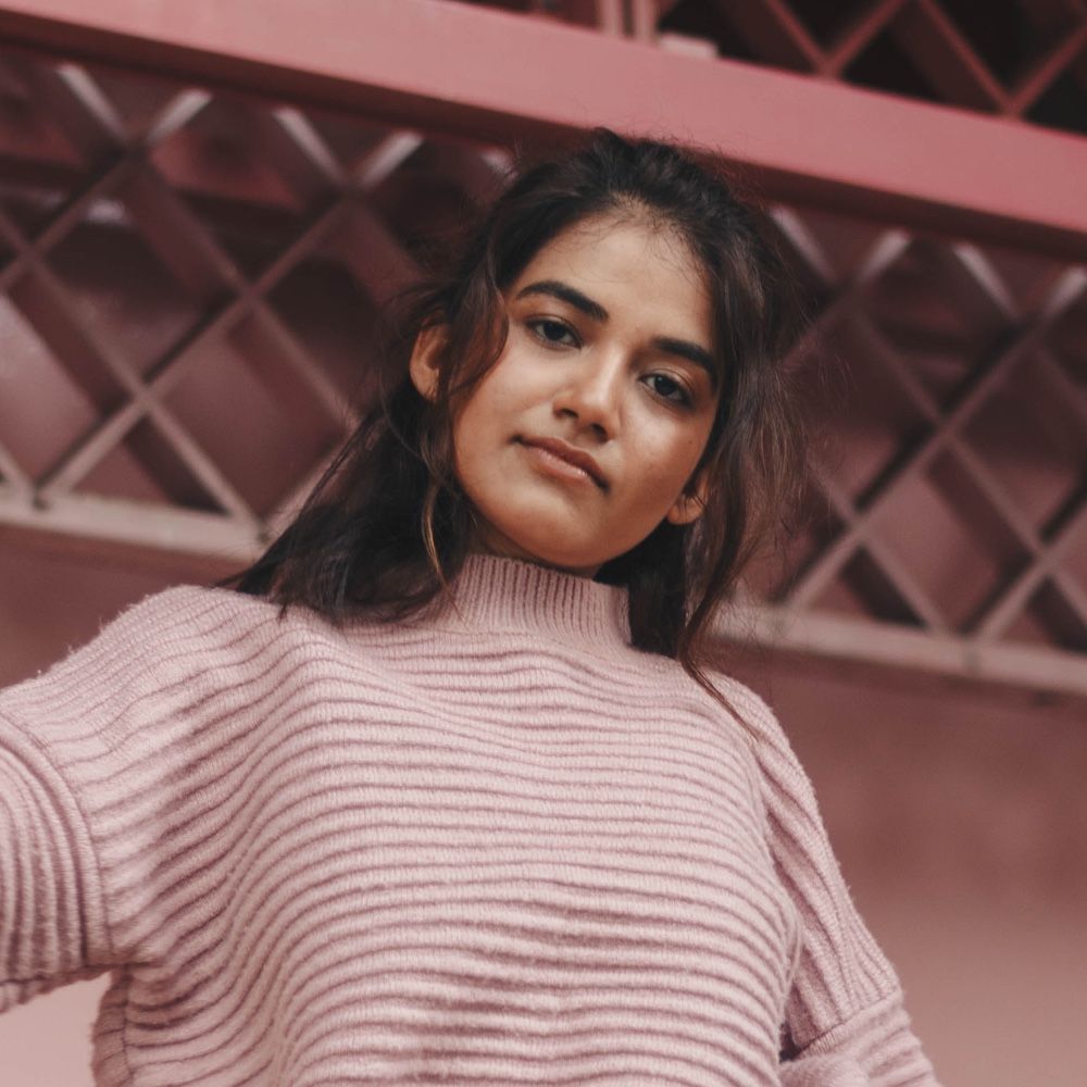 A woman in a pink sweater is standing in front of a pink wall.