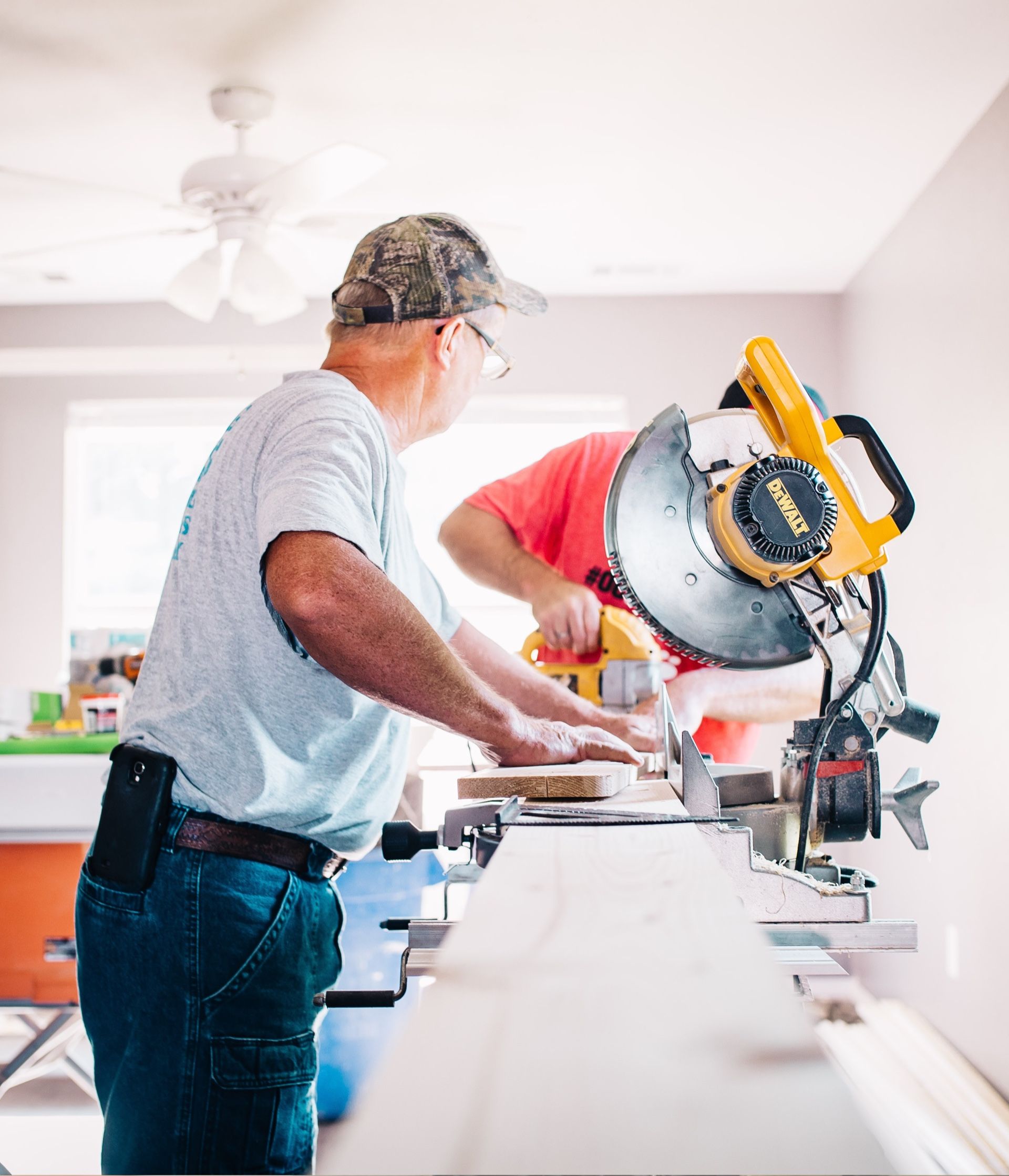 Two people using a miter saw to cut wood indoors. One wears a camo hat, the other a pink shirt.