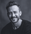 Man with dark hair and beard smiles at the camera. Black and white portrait.