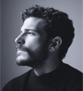 Man with dark curly hair and beard looks to the right, wearing a black turtleneck.