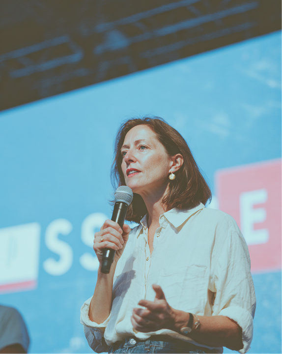Woman speaking into a microphone, gesturing with hands. White shirt, blue background.