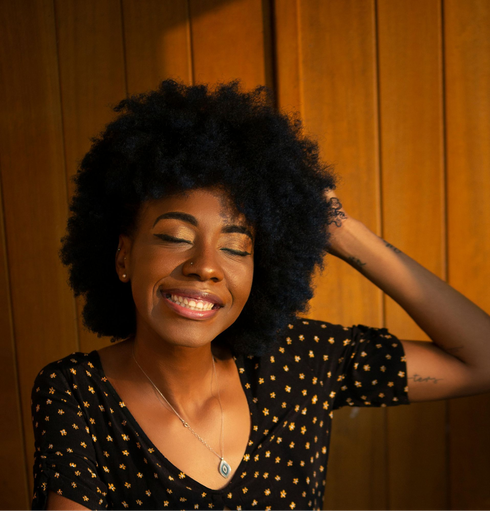 Smiling woman with curly hair in a black polka-dot top, posing against a wooden wall.