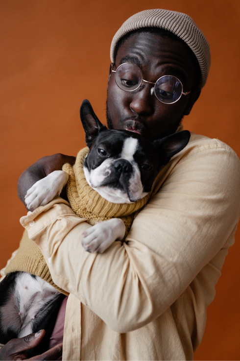 Person in beige sweater holding a black-and-white dog wearing a yellow knit sweater against an orange background