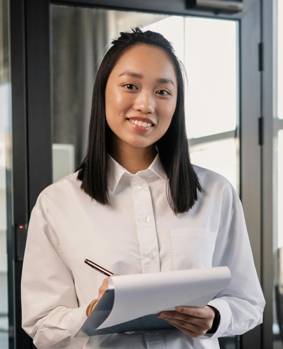 Woman in white shirt, holding a clipboard and pen, smiling in front of a glass door.