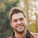Man smiling, outdoors; brown leather jacket, blurred autumn background.