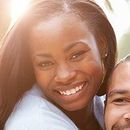 Woman smiling, light background, shoulder and eye of another person visible.