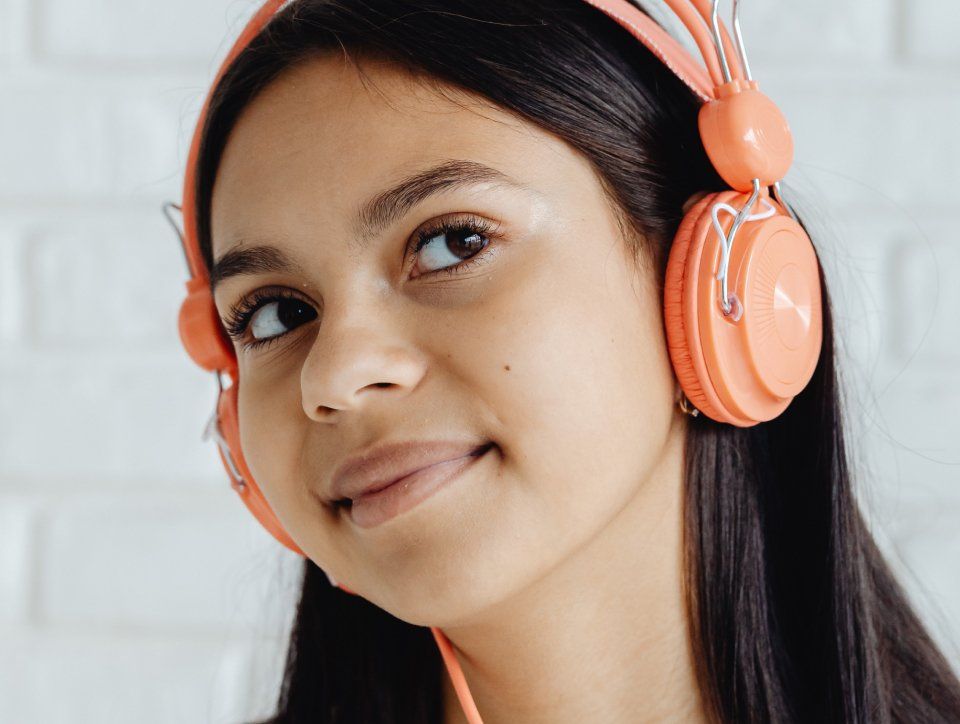 Person wearing coral headphones smiles, looking up and left against a white brick wall.