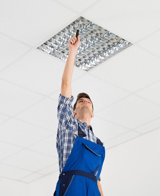 Man in blue overalls reaching up toward a ceiling light fixture in a white-tiled ceiling.