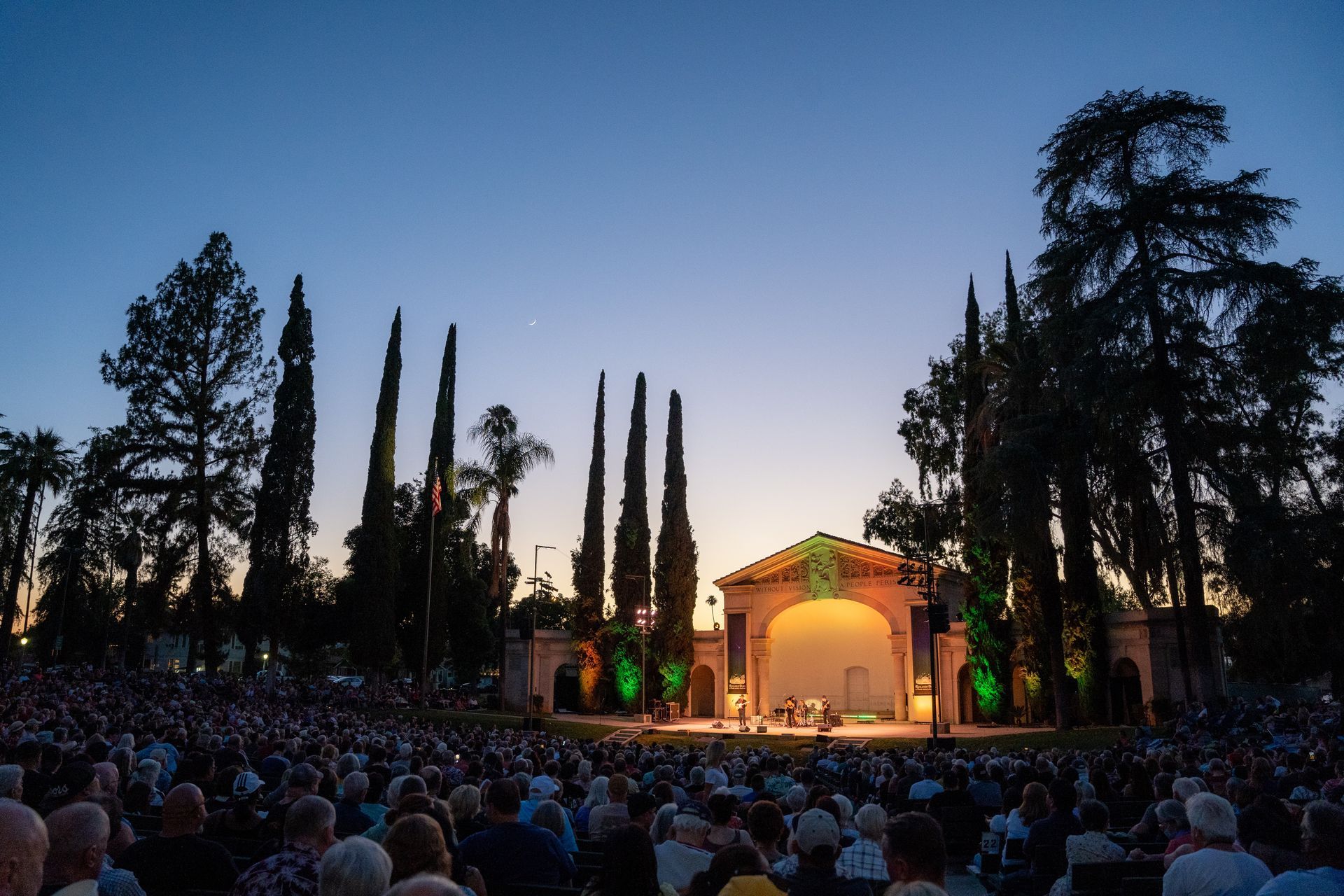 Outdoor concert at dusk: stage with performers, large crowd seated, tall trees, arched structure, and a colorful sky.