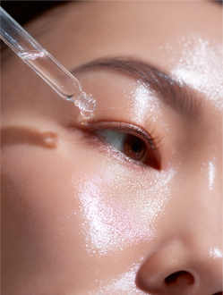 A clear dropper applies a droplet of serum to a person's glowing, hydrated cheek near their eye.