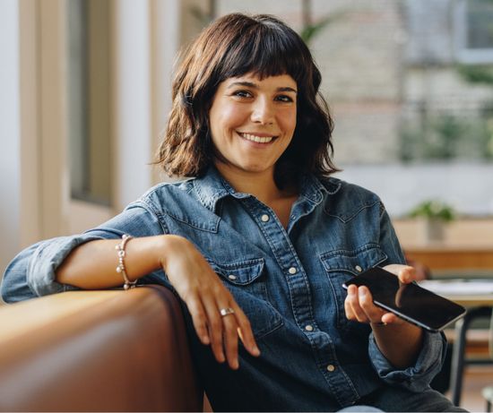 Woman with dark hair smiles, holding a phone, sitting on a sofa in a brightly lit room.