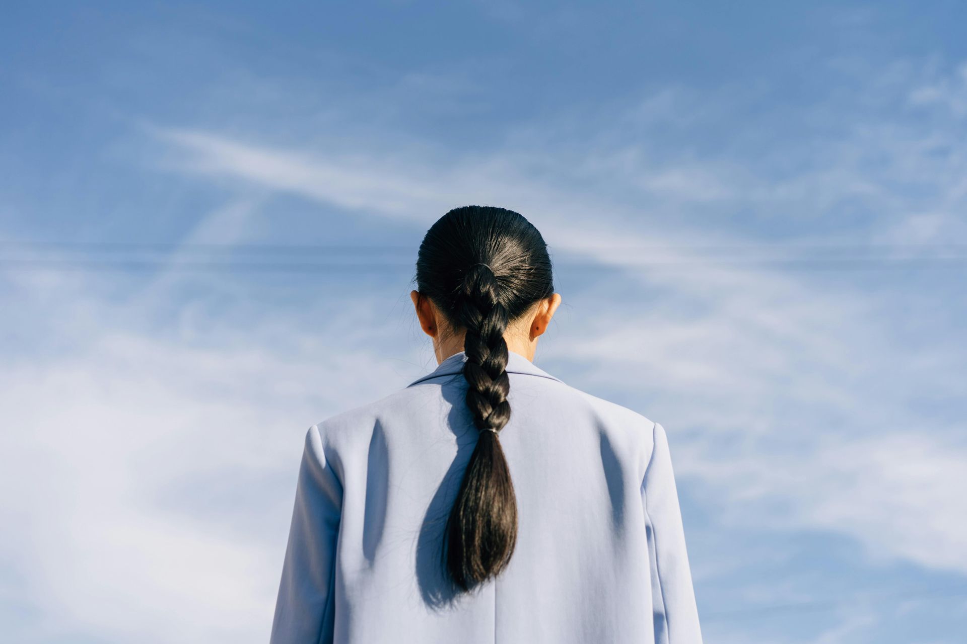 Person in blue blazer, back to the camera, with a braid against a blue sky.