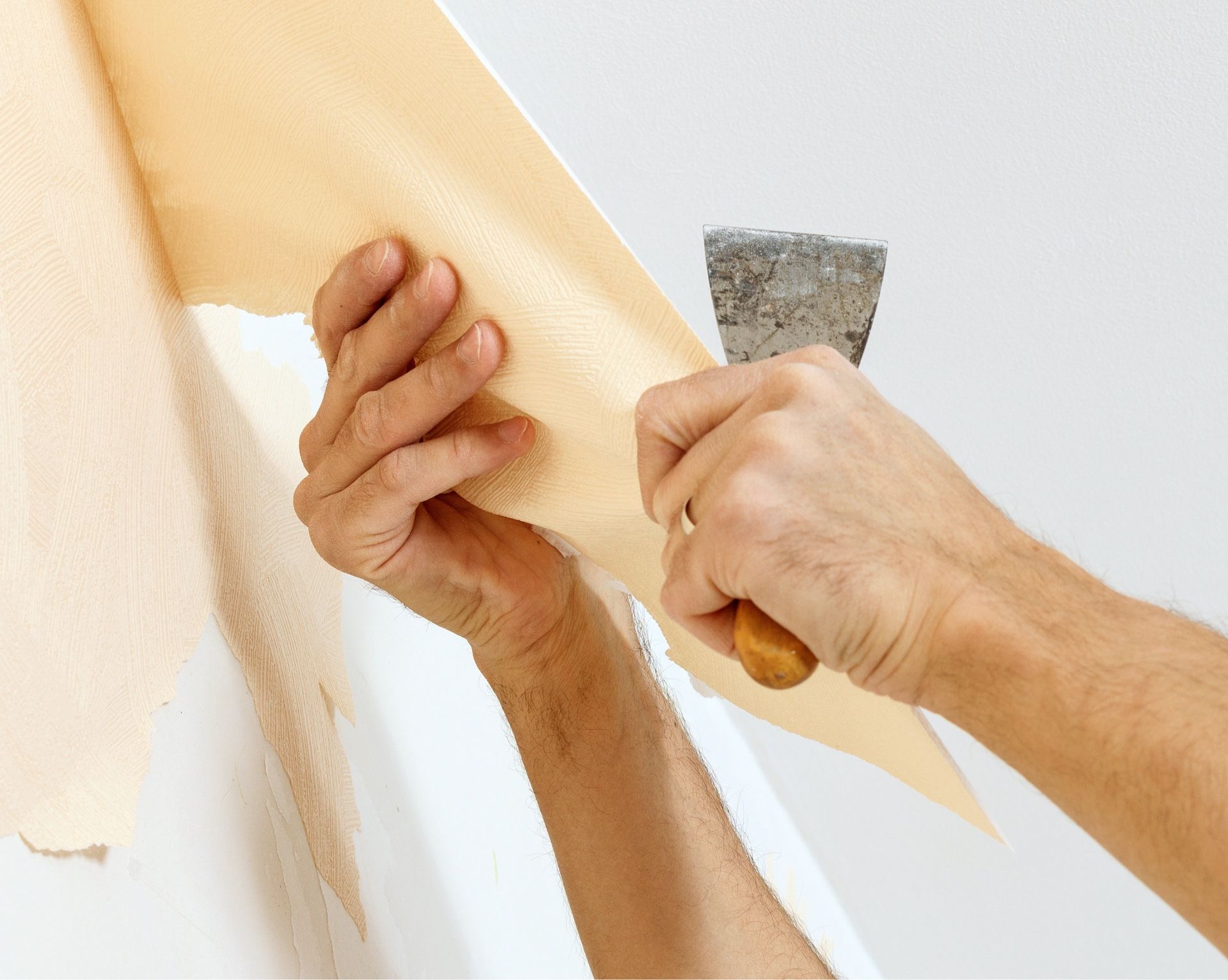 Hands peeling off old wallpaper from a white wall with a putty knife.