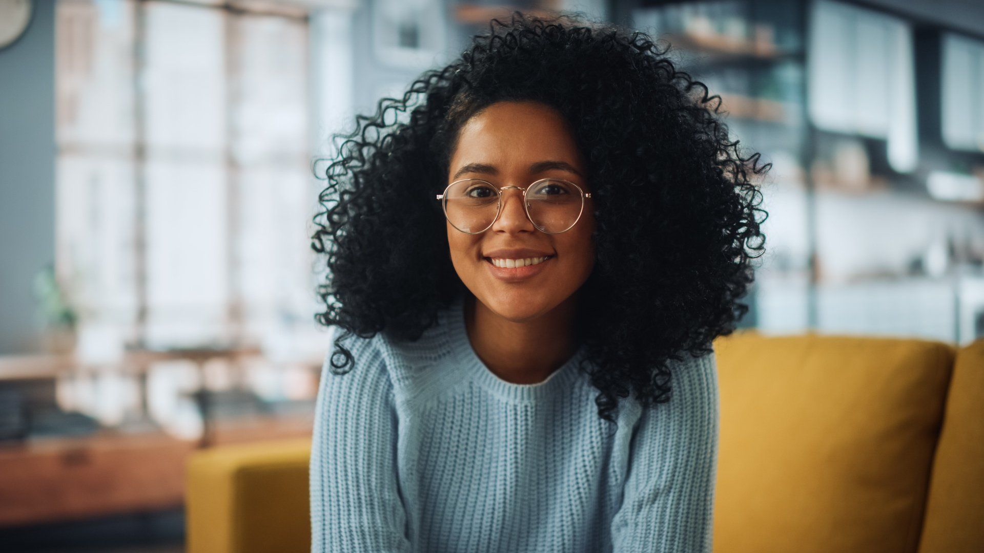 A woman wearing glasses and a blue sweater is sitting on a yellow couch.