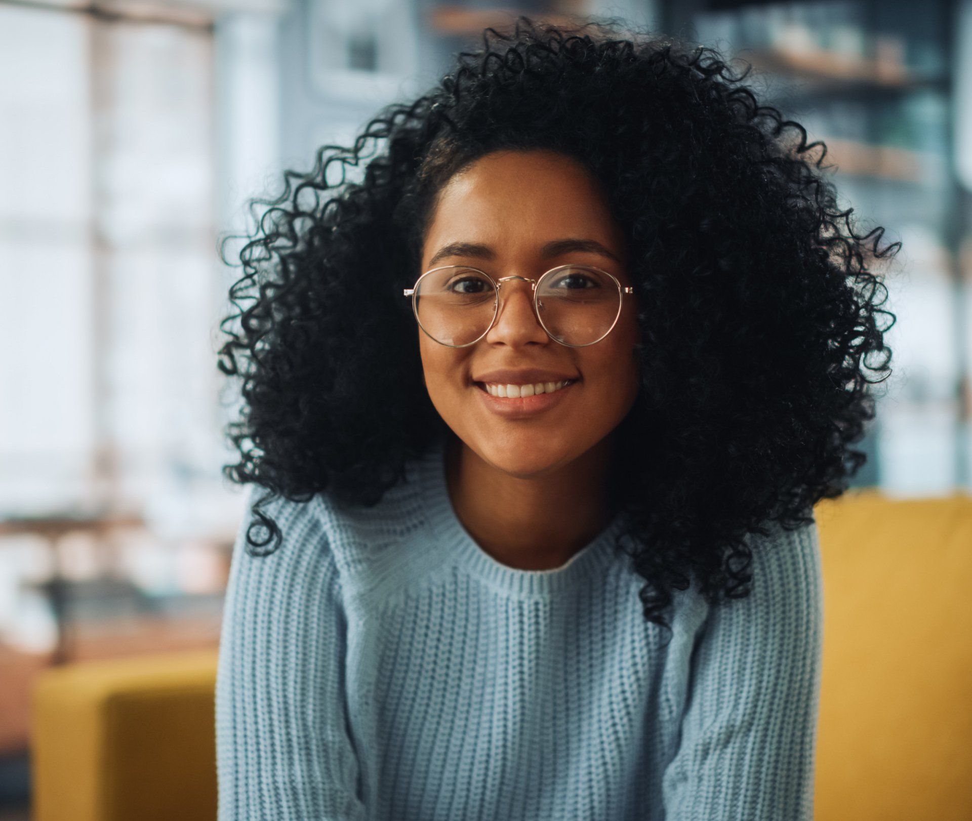 A woman with curly hair is wearing glasses and a blue sweater.