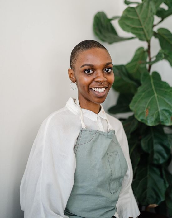 A woman wearing an apron and a white shirt is smiling in front of a plant.