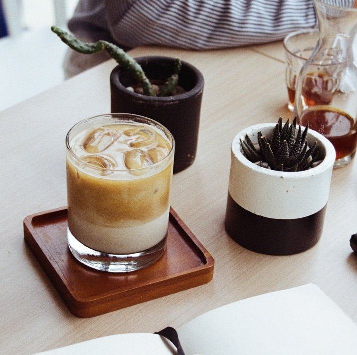 A glass of iced coffee sits on a wooden coaster next to a potted plant