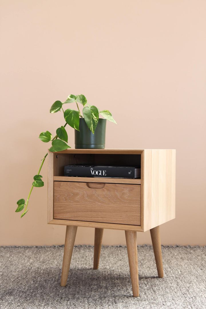Mesita de noche de madera con planta y cajón, apoyada contra una pared color melocotón, sobre una alfombra gris.