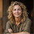 Smiling woman with curly blonde hair in an olive shirt, arms crossed, against a dark background